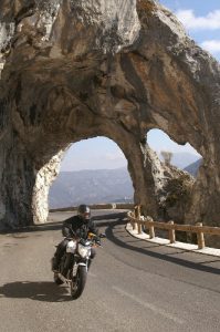 A motorbike with a rider driving on a mountain road just passed an arch made of rock.